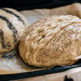 Två bröd ligger på bakplåtspapper i nedre delen av ugnen. Ett är toppat med vallmofrön, det andra med sesamfrön och en flätad degdekoration.