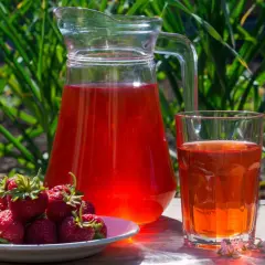 En karaff och ett glas med röd saft står på ett träbord. En tallrik med färska jordgubbar bredvid. Perfekt för ett enkelt jordgubbssaft recept.