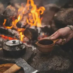 En hand håller en träkopp med kaffe vid en lägereld. En kaffepanna och en yxa ligger bredvid. Perfekt kokkaffe dosering för en mysig stund.