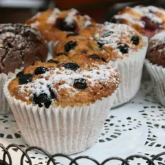 Flera vanliga muffins, några med blåbär och pudrade med florsocker, ligger på ett vitt spetsfat.