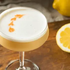 En läcker drink med skummigt toppskikt, garnerad med apelsin. Perfekt för ett sifon recept. Citron och drinkverktyg i bakgrunden.