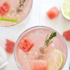 Två glas med en rosa, alkoholfri drink med is, vattenmelonbitar, lime och rosmarin.
