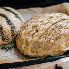 Två bröd ligger på bakplåtspapper i nedre delen av ugnen. Ett är toppat med vallmofrön, det andra med sesamfrön och en flätad degdekoration.