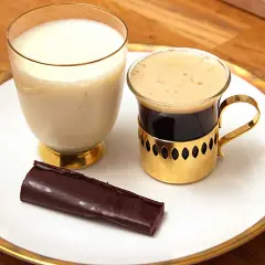 En bit mörk choklad ganache ligger bredvid en kopp kaffe och ett glas mjölk på en vit tallrik med guldkant.