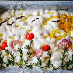 Focaccia surdeg dekorerad som en sommaräng med tomater, rödlök och rosmarin. Fåglar flyger över en sol av gula tomater.