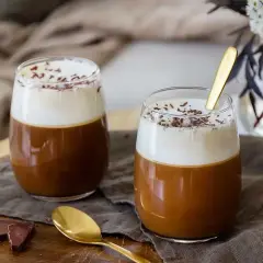 Två glas med krämig kaffe Karlsson, toppad med vispgrädde och chokladströssel. En guldig sked vilar i det ena glaset.