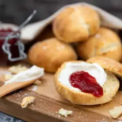 Nybakade engelska scones med sylt och grädde på en träskärbräda. En burk sylt syns i bakgrunden.