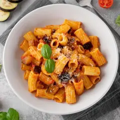 En skål med rig pasta alla norma, toppad med basilika och riven ost. Bredvid syns aubergineskivor, körsbärstomater och färsk basilika.