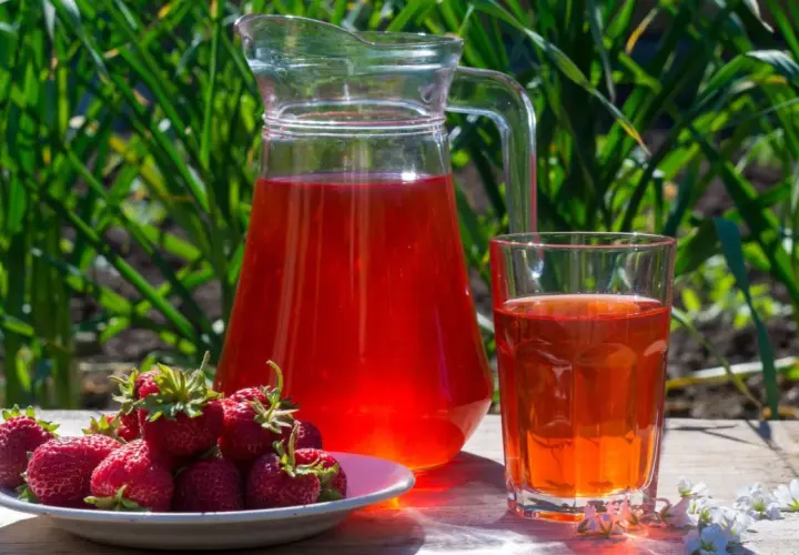 En karaff och ett glas med röd saft står på ett träbord. En tallrik med färska jordgubbar bredvid. Perfekt för ett enkelt jordgubbssaft recept.