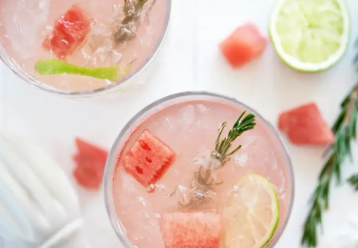 Två glas med en rosa, alkoholfri drink med is, vattenmelonbitar, lime och rosmarin.