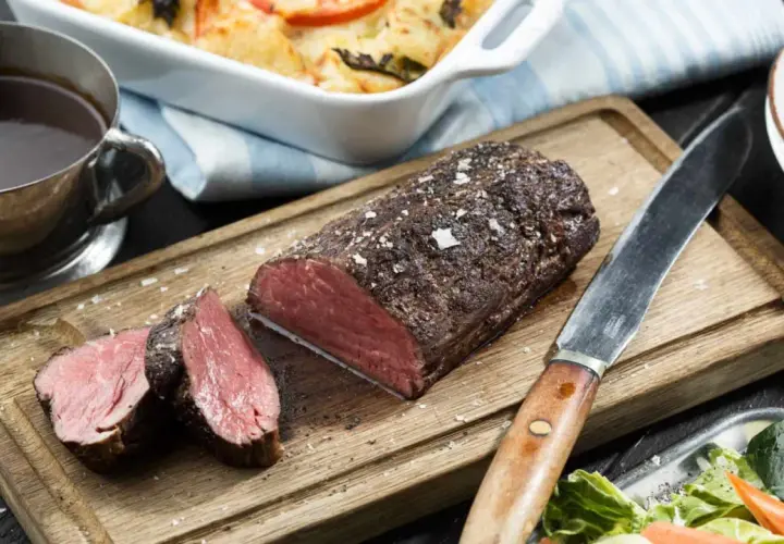 En saftig, helstekt oxfile med perfekt rosa kärna, serverad på en träskärbräda med en skål fylld med mörk sås och en fräsch sallad.