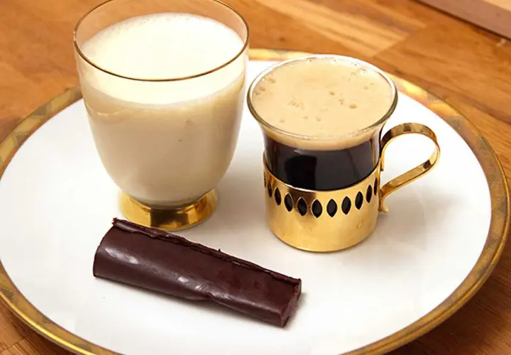En bit mörk choklad ganache ligger bredvid en kopp kaffe och ett glas mjölk på en vit tallrik med guldkant.