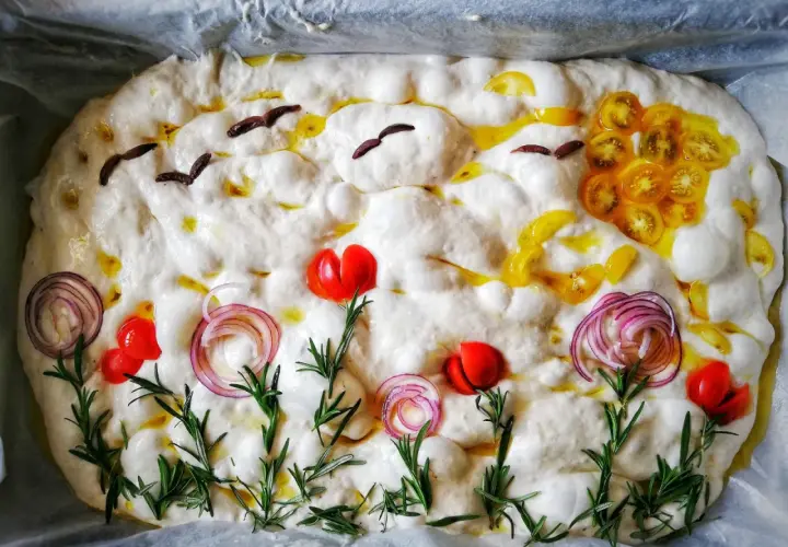 Focaccia surdeg dekorerad som en sommaräng med tomater, rödlök och rosmarin. Fåglar flyger över en sol av gula tomater.