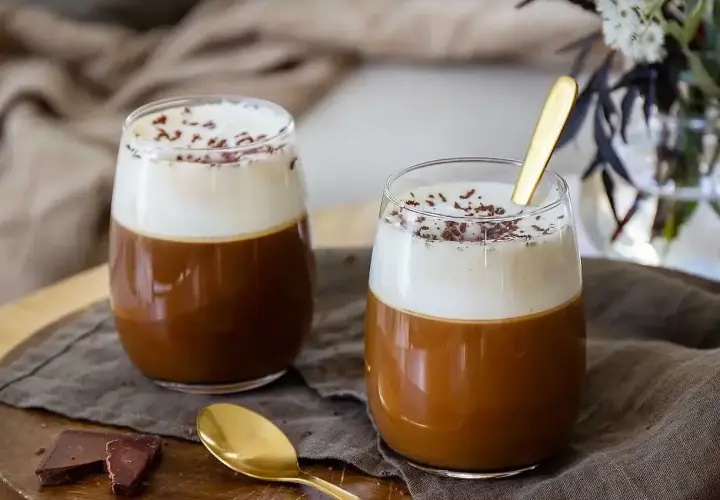 Två glas med krämig kaffe Karlsson, toppad med vispgrädde och chokladströssel. En guldig sked vilar i det ena glaset.