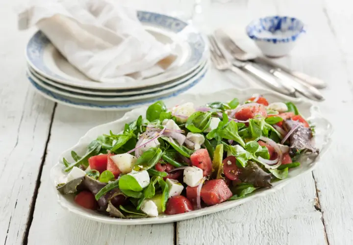 En uppfriskande melonsallad med fetaost, färska gröna blad, tomater och rödlök, ringlad med olivolja. Perfekt sommarbuffé!
