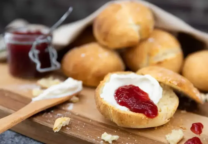 Nybakade engelska scones med sylt och grädde på en träskärbräda. En burk sylt syns i bakgrunden.