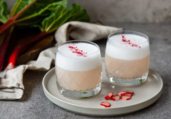 Två glas med en krämig rabarber sour, toppad med skum och strödd med rabarberpulver. En härlig sommardrink!