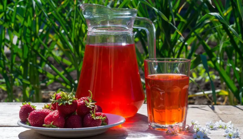 En karaff och ett glas med röd saft står på ett träbord. En tallrik med färska jordgubbar bredvid. Perfekt för ett enkelt jordgubbssaft recept.