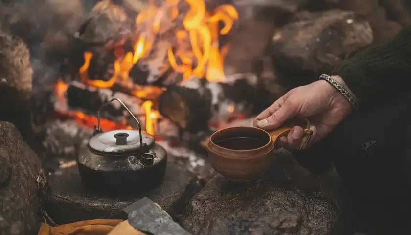 En hand håller en träkopp med kaffe vid en lägereld. En kaffepanna och en yxa ligger bredvid. Perfekt kokkaffe dosering för en mysig stund.