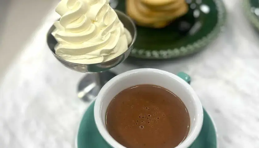 En kopp varm choklad med vispgrädde bredvid. En stapel pannkakor med blåbär syns i bakgrunden.