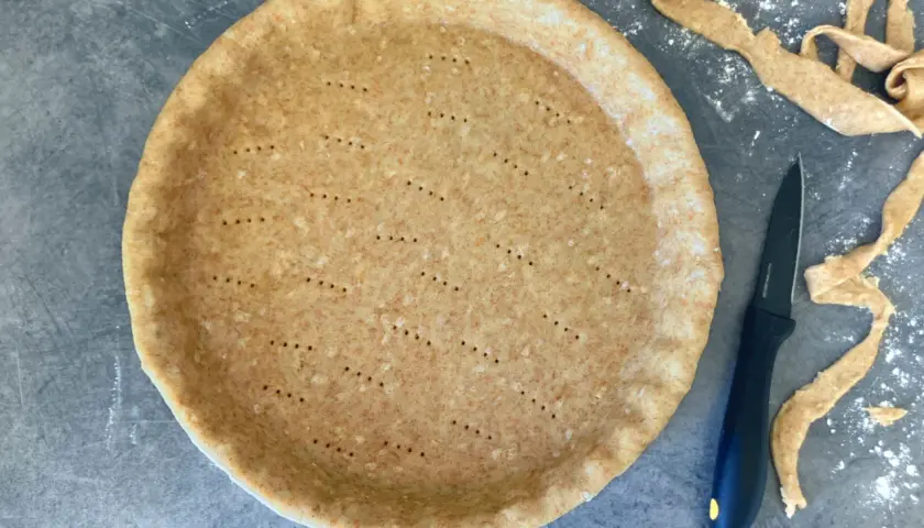 En förberedd pajdeg med prickar, redo för ett paj recept. Bredvid ligger degrester och en kniv.