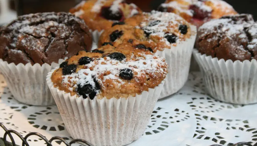 Flera vanliga muffins, några med blåbär och pudrade med florsocker, ligger på ett vitt spetsfat.