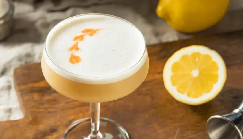 En läcker drink med skummigt toppskikt, garnerad med apelsin. Perfekt för ett sifon recept. Citron och drinkverktyg i bakgrunden.