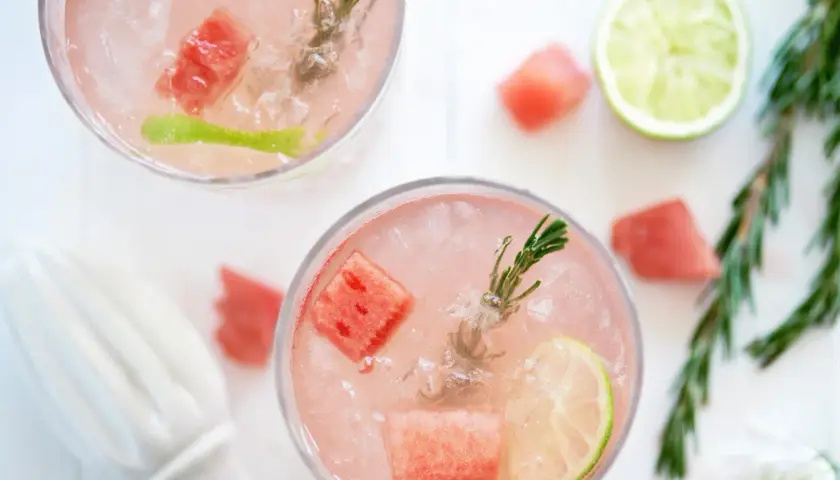 Två glas med en rosa, alkoholfri drink med is, vattenmelonbitar, lime och rosmarin.