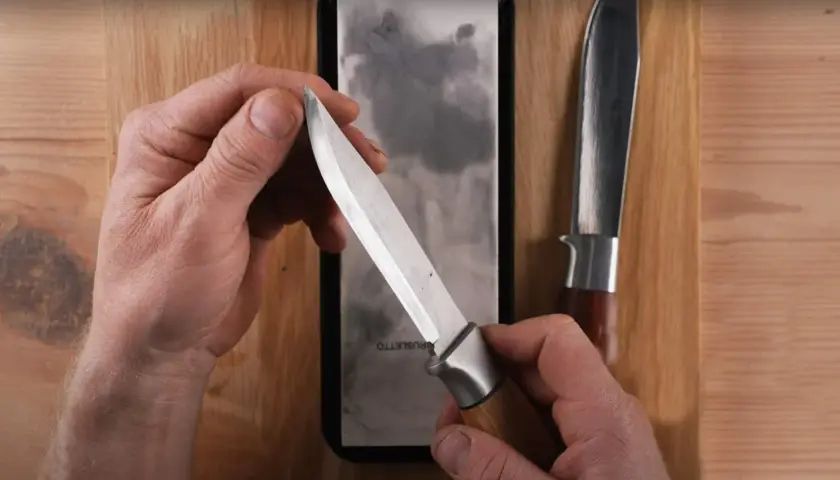 Händer som håller en kniv över en slipsten, visar hur slipar man knivar för att få en vass egg. En annan kniv ligger bredvid.