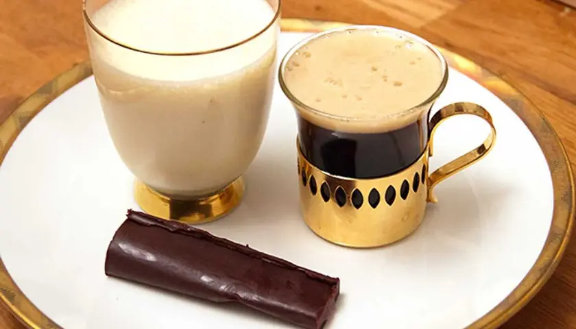 En bit mörk choklad ganache ligger bredvid en kopp kaffe och ett glas mjölk på en vit tallrik med guldkant.