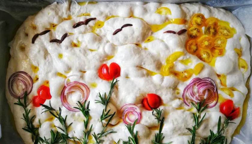 Focaccia surdeg dekorerad som en sommaräng med tomater, rödlök och rosmarin. Fåglar flyger över en sol av gula tomater.