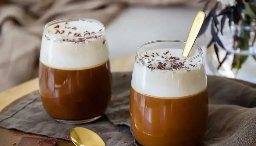 Två glas med krämig kaffe Karlsson, toppad med vispgrädde och chokladströssel. En guldig sked vilar i det ena glaset.