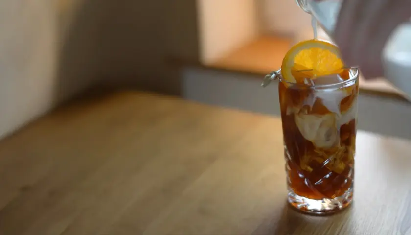 En hand häller mjölk i ett glas med iskaffe, garnerat med en apelsinskiva. Ett perfekt iskaffe recept för en varm dag.