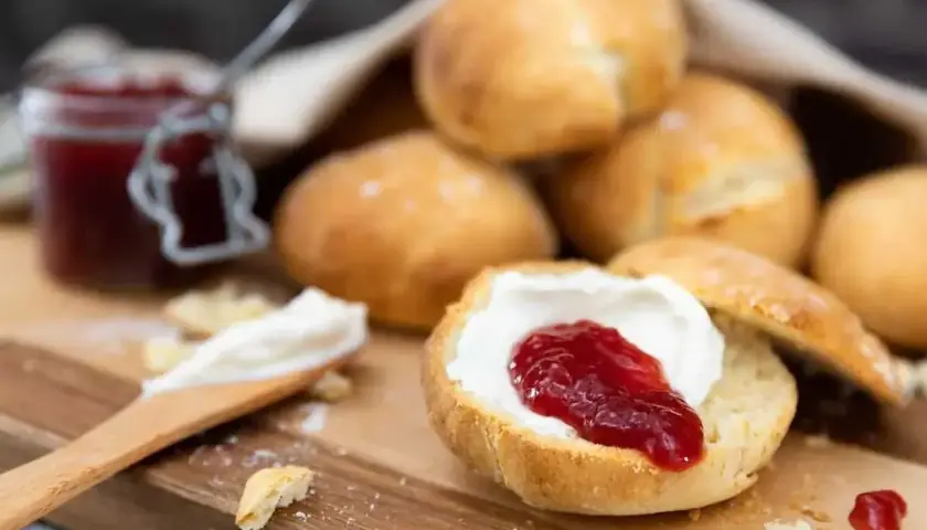 Nybakade engelska scones med sylt och grädde på en träskärbräda. En burk sylt syns i bakgrunden.