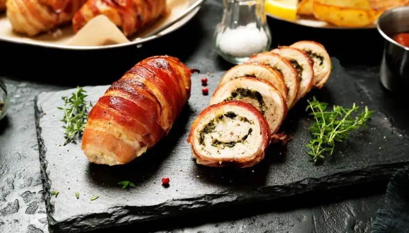 En hel och skivad baconlindad kyckling med örter, serverad på en svart stenplatta med timjan.