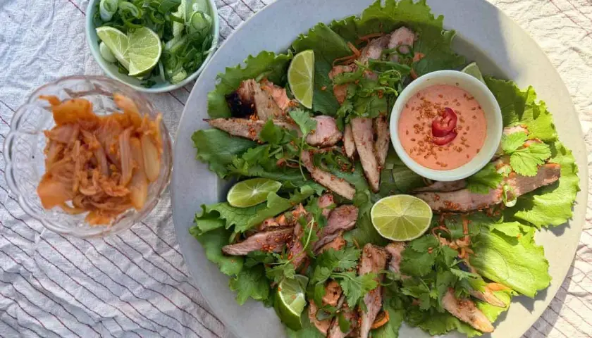 Grilla pluma serveras i salladsblad med lime, koriander och en krämig dipp. Kimchi och salladslök i skålar bredvid.