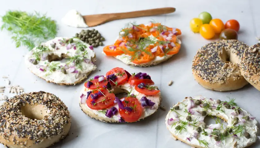 Flera bröd med hål i mitten, toppade med färskost, lax, tomater, rödlök och dill. Ett härligt urval av bagels!