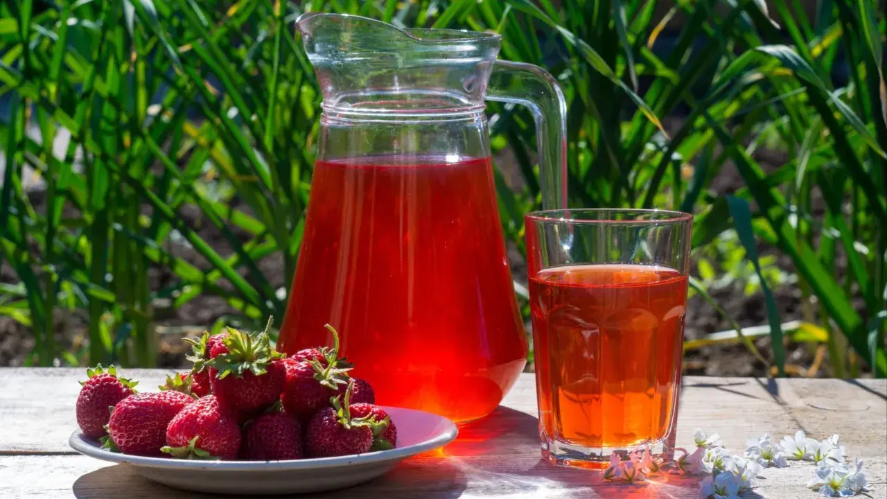 En karaff och ett glas med röd saft står på ett träbord. En tallrik med färska jordgubbar bredvid. Perfekt för ett enkelt jordgubbssaft recept.