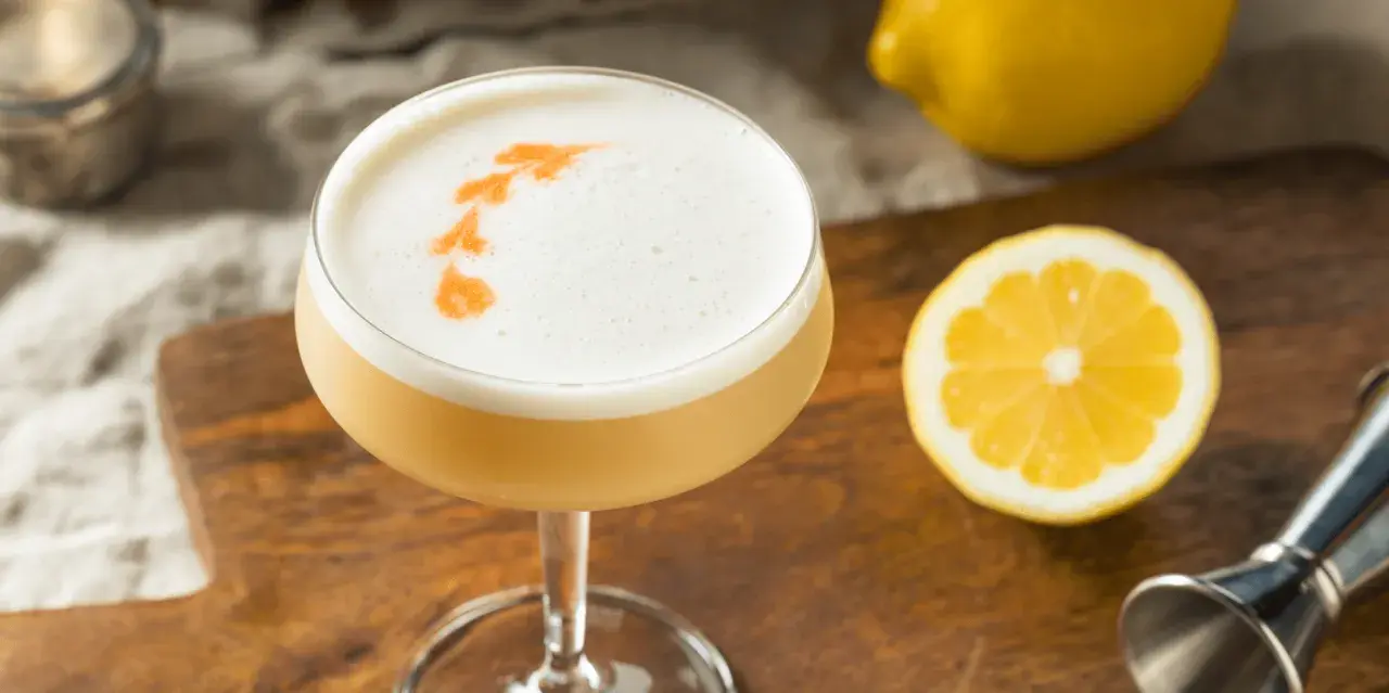En läcker drink med skummigt toppskikt, garnerad med apelsin. Perfekt för ett sifon recept. Citron och drinkverktyg i bakgrunden.