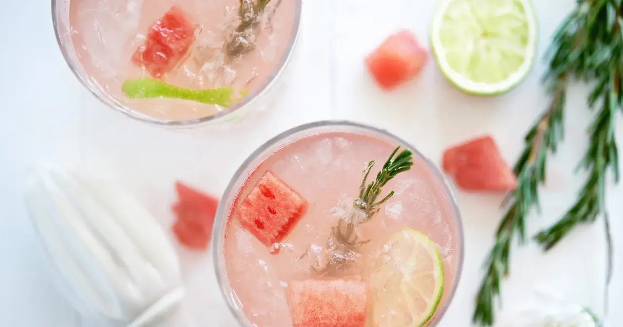 Två glas med en rosa, alkoholfri drink med is, vattenmelonbitar, lime och rosmarin.