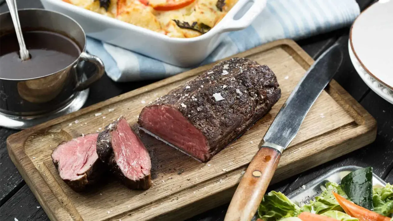 En saftig, helstekt oxfile med perfekt rosa kärna, serverad på en träskärbräda med en skål fylld med mörk sås och en fräsch sallad.