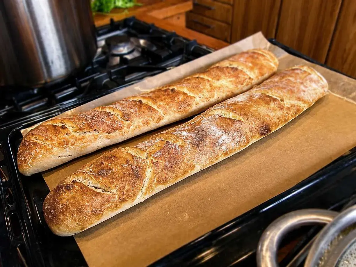 Två gyllenbruna surdegsbaguetter på bakplåtspapper, redo att avnjutas. Ett perfekt surdegsbaguette recept!