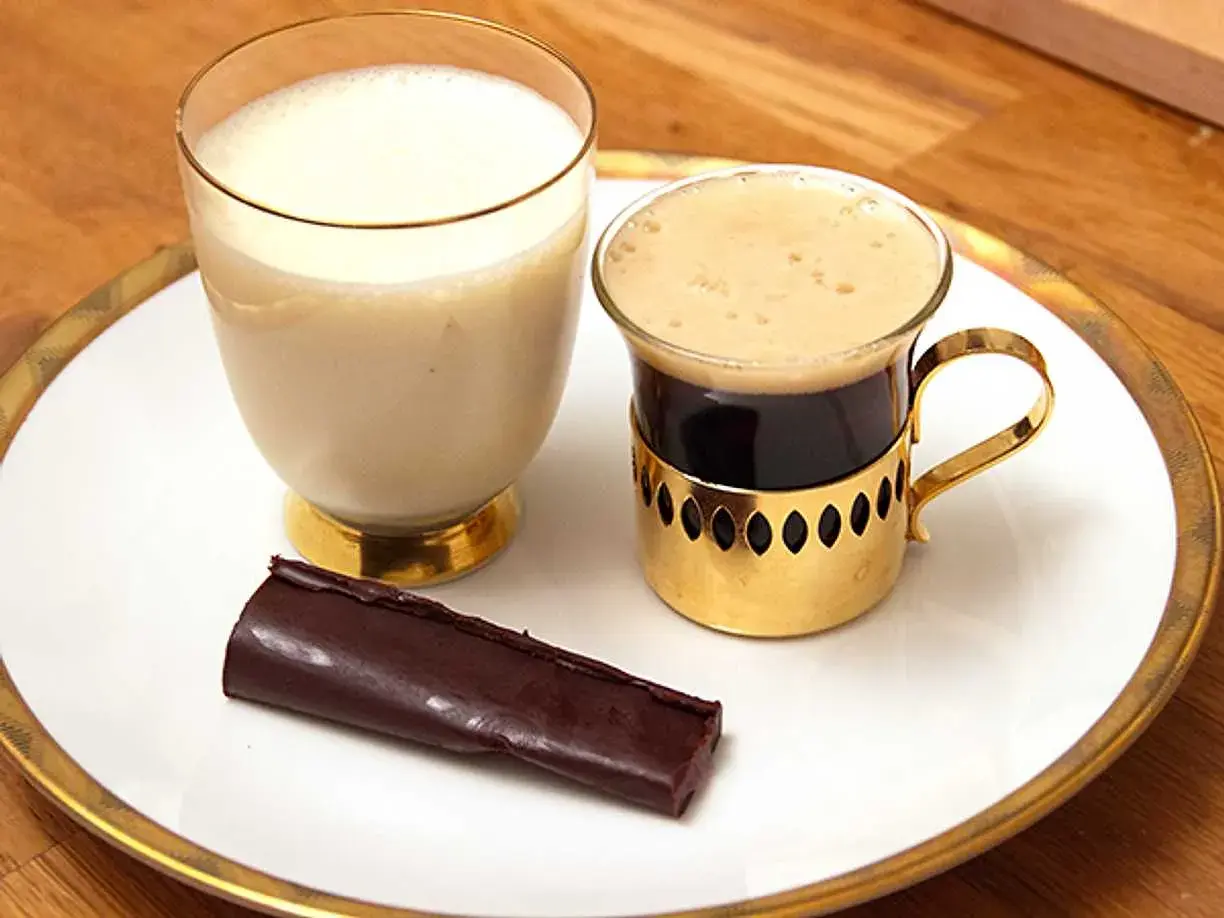 En bit mörk choklad ganache ligger bredvid en kopp kaffe och ett glas mjölk på en vit tallrik med guldkant.