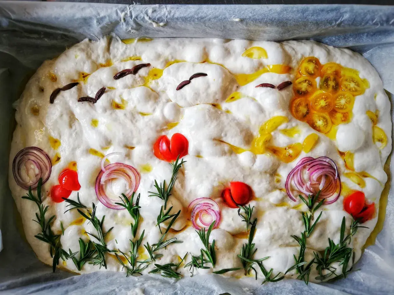 Focaccia surdeg dekorerad som en sommaräng med tomater, rödlök och rosmarin. Fåglar flyger över en sol av gula tomater.