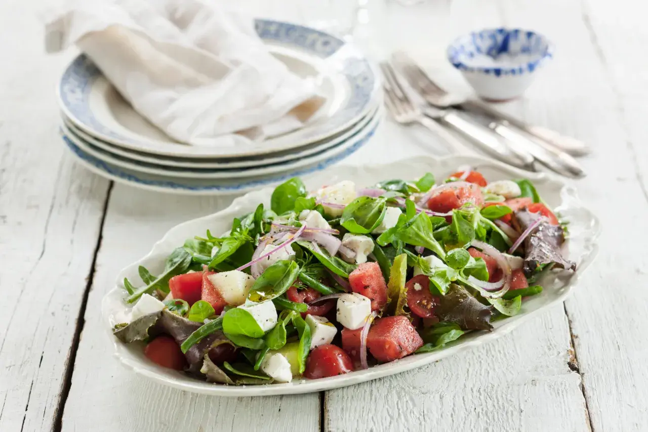 En uppfriskande melonsallad med fetaost, färska gröna blad, tomater och rödlök, ringlad med olivolja. Perfekt sommarbuffé!