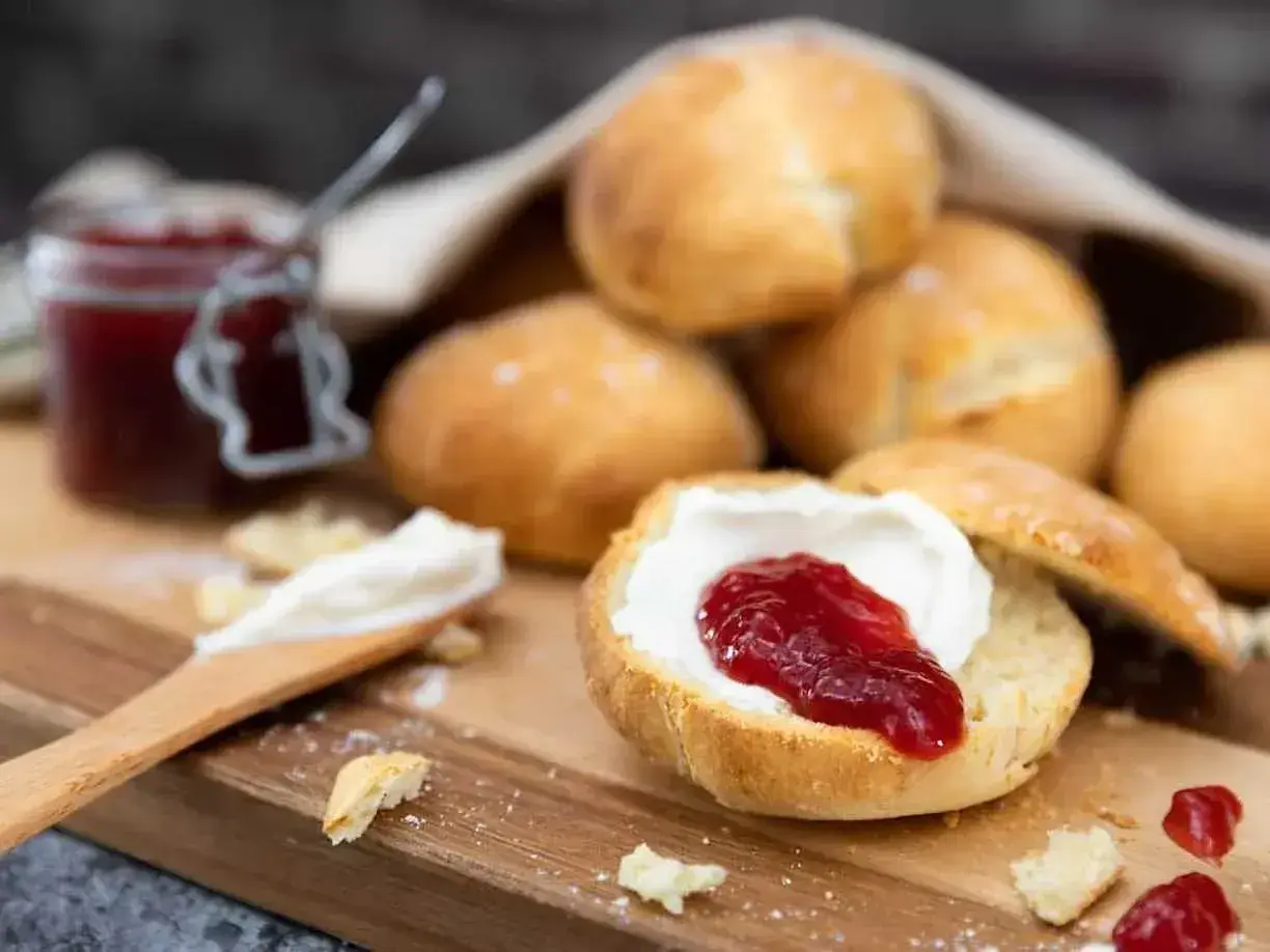 Nybakade engelska scones med sylt och grädde på en träskärbräda. En burk sylt syns i bakgrunden.