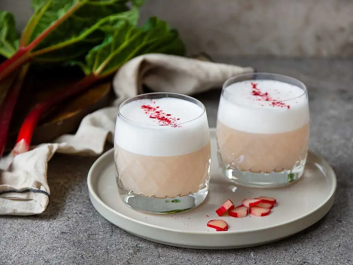 Två glas med en krämig rabarber sour, toppad med skum och strödd med rabarberpulver. En härlig sommardrink!