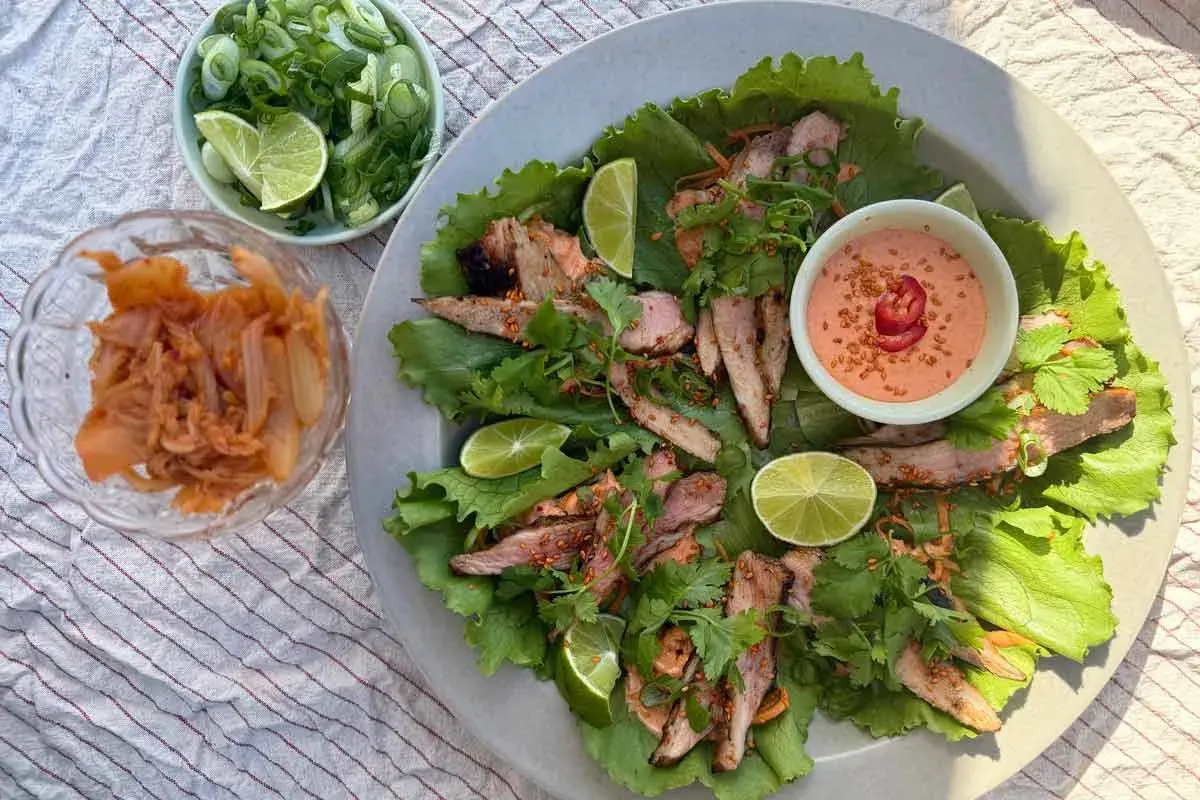 Grilla pluma serveras i salladsblad med lime, koriander och en krämig dipp. Kimchi och salladslök i skålar bredvid.