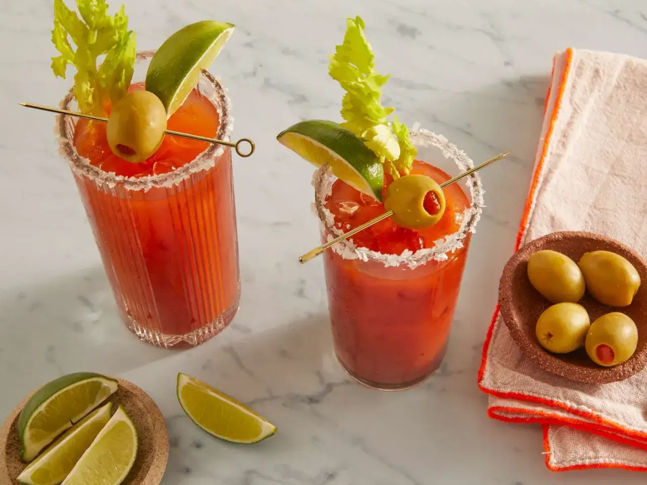 Tv&aring; Bloody Mary-drinkar med saltkant, lime, oliv och selleri. En sk&aring;l med oliver och limeklyftor ligger bredvid.