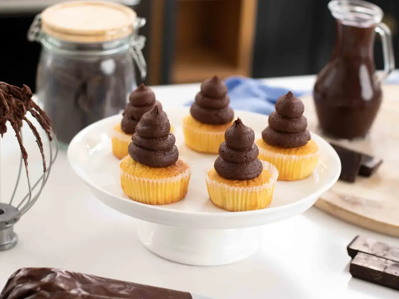Små cupcakes med chokladfrosting, perfekt för ett ganache recept till praliner. En glasburk med choklad och en kanna med chokladsås i bakgrunden.